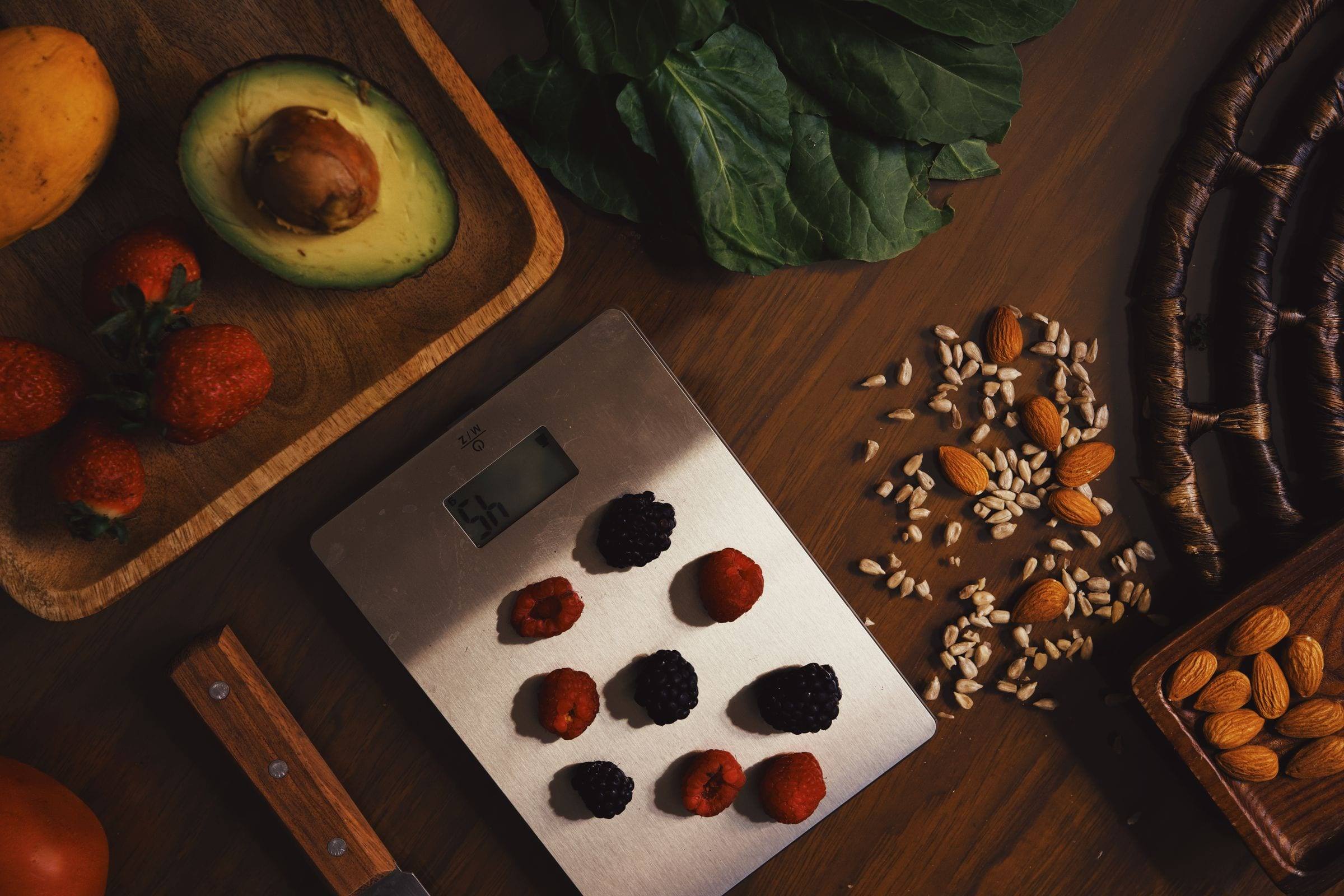 Küchenwaage mit Beeren, daneben Avocado, Mandeln und Kerne auf einem Holzbrett