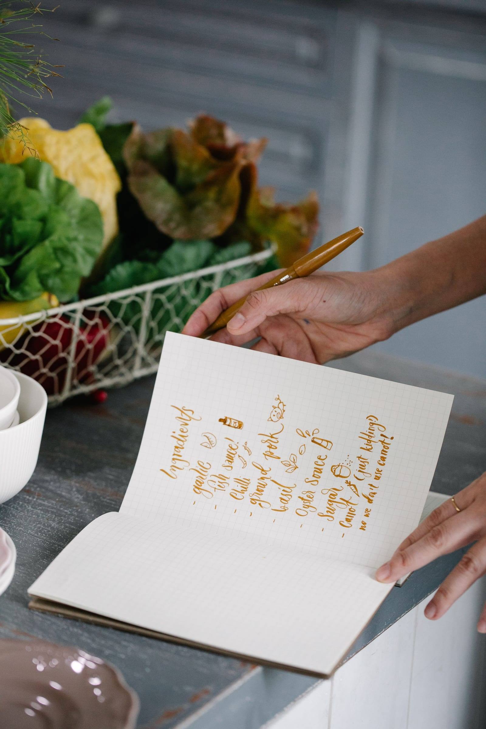Hand notiert mit einem goldenen Stift Zutaten in ein Heft, dahinter frisches Gemüse