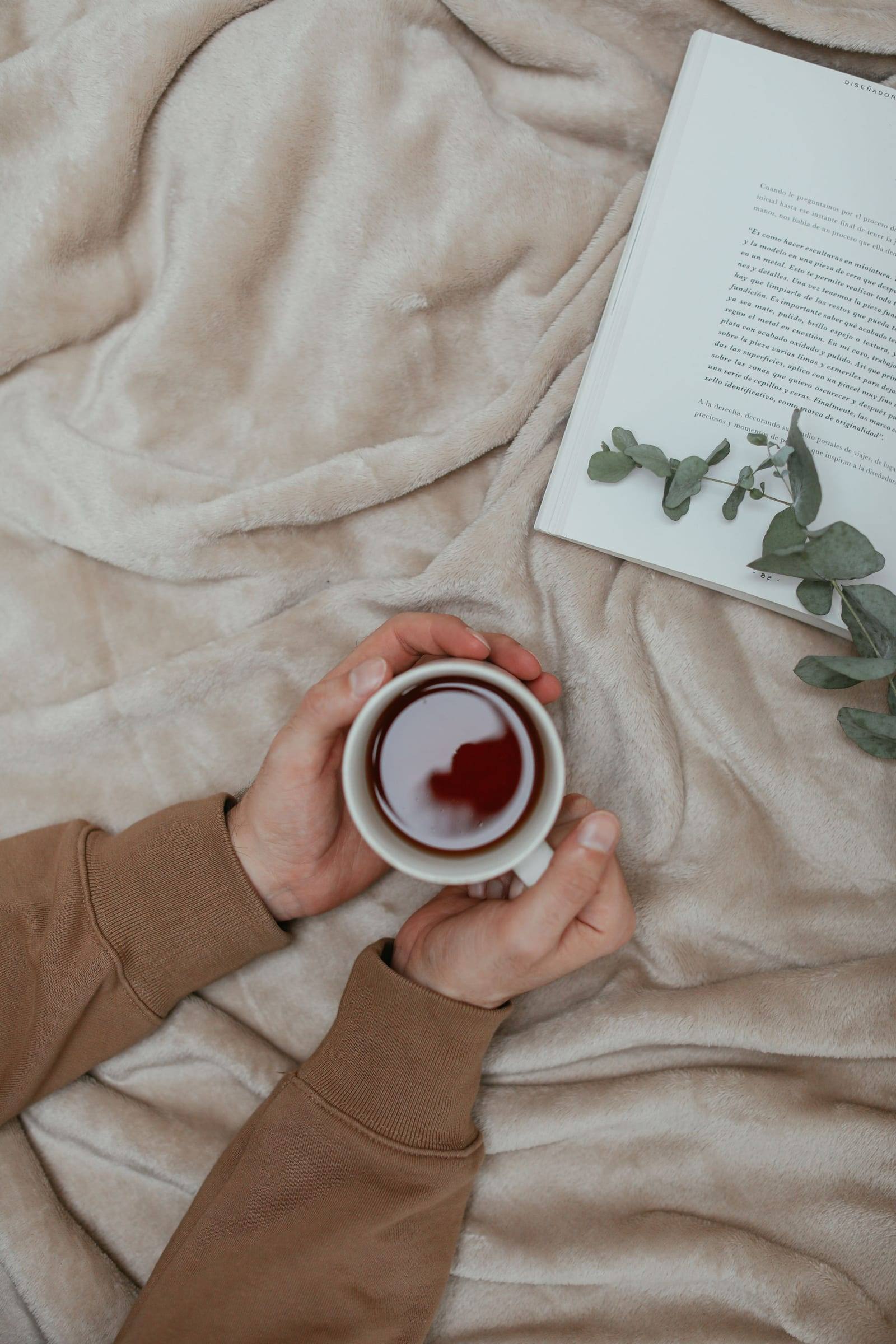 Hände halten eine Tasse Tee auf einer hellen Wolldecke, daneben ein aufgeschlagenes Buch