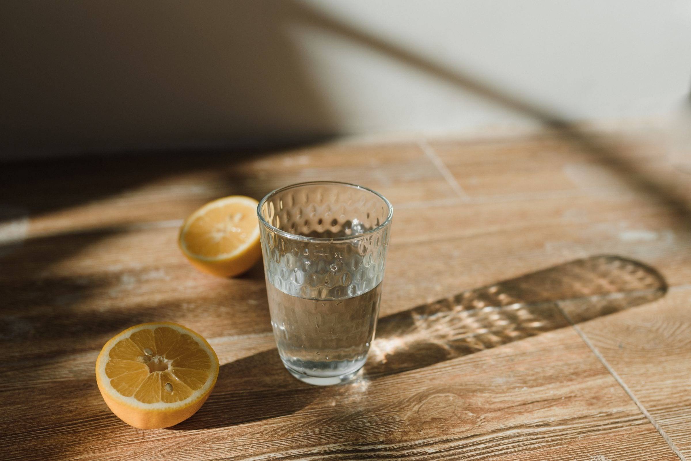 Glas Wasser neben halbierten Zitronen auf einem Holztisch im Sonnenlicht